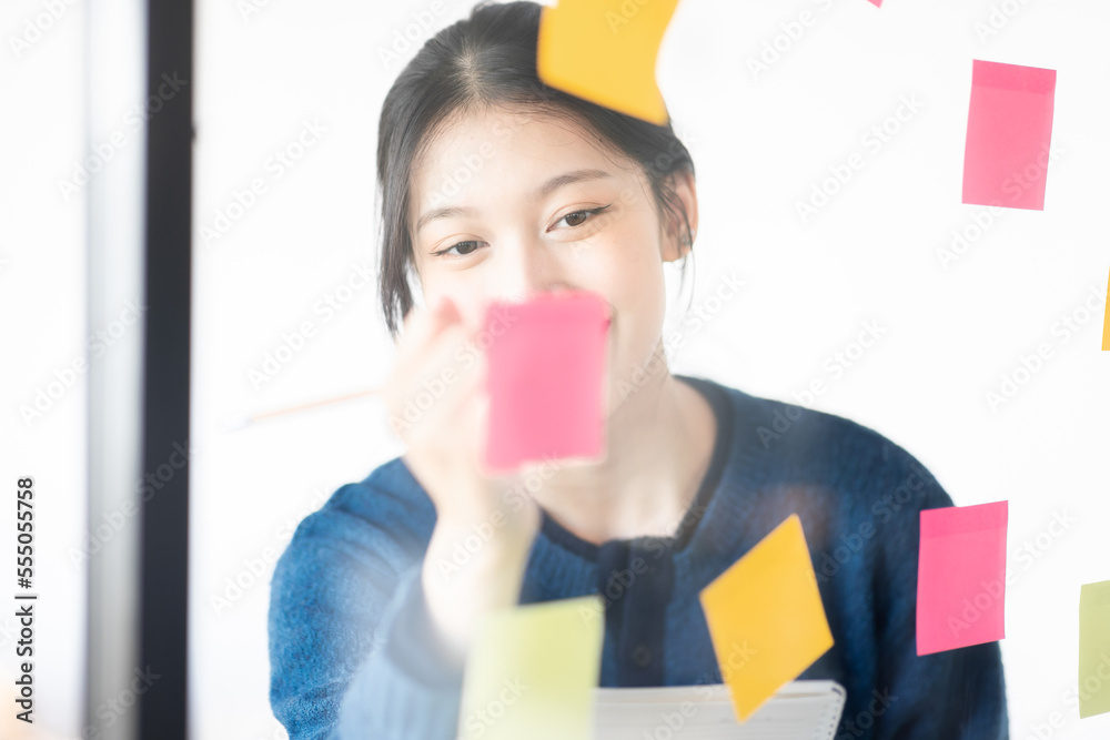Portrait shot of Cheerful young adult business Asian woman sticking adhesive paper notes to a glass wall, idea-sharing board in business office concept. Portrait shot of Cheerful young adult business Asian woman sticking adhesive paper notes to a glass wall, idea-sharing board in business office concept.