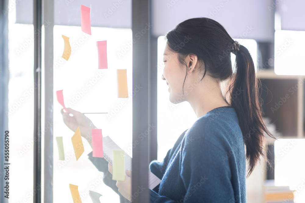 Portrait shot of Cheerful young adult business Asian woman sticking adhesive paper notes to a glass wall, idea-sharing board in business office concept. Portrait shot of Cheerful young adult business Asian woman sticking adhesive paper notes to a glass wall, idea-sharing board in business office concept.