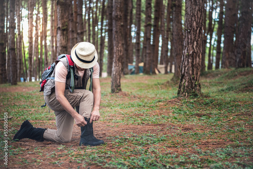 Asian male tourist sitting under a pine tree and wearing boots to prepare for travel in the forest. to backpack and travel concept.