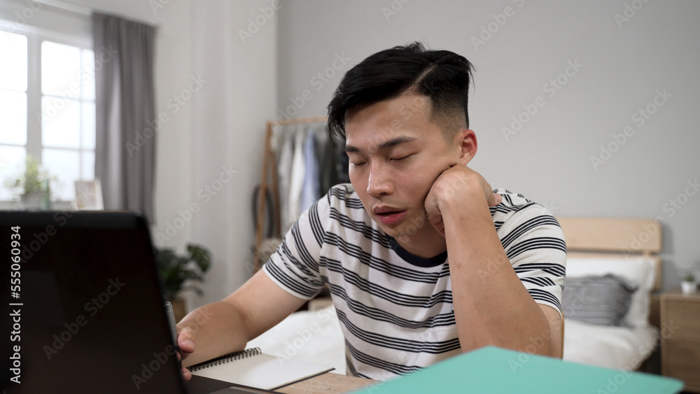 sleepy asian male student learning from home is propping face and nodding off while taking online course on the computer in the bedroom during quarantine. sleepy asian male student learning from home is propping face and nodding off while taking online course on the computer in the bedroom during quarantine.