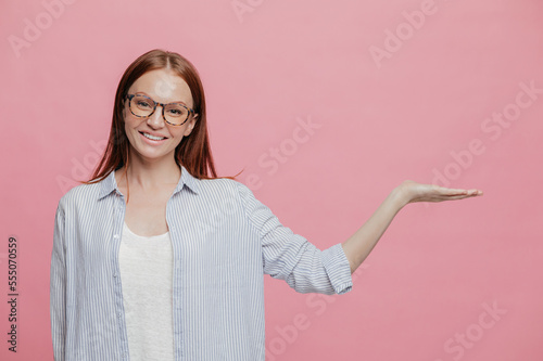 Positive young smiling woman raises hand, pretends holding something, gestures over copy space, dressed in sylish oversized shirt, wears spectacles, isolated over pink studio wall. Advertisement