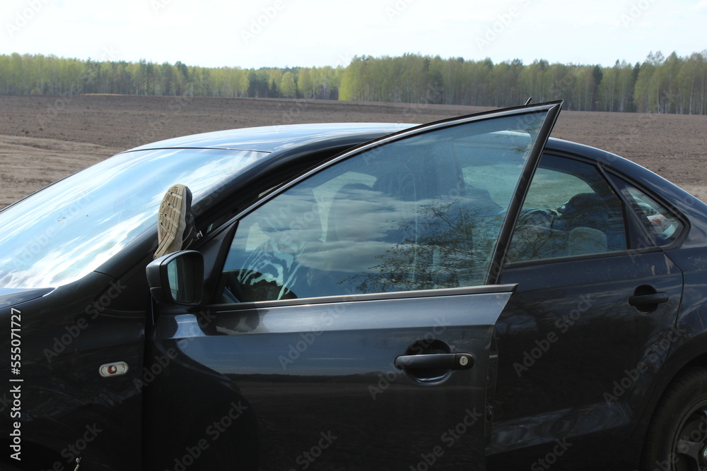 the girl is sitting in the driver's seat in the car the door is ajar the leg is lying on the door the man behind the glass
