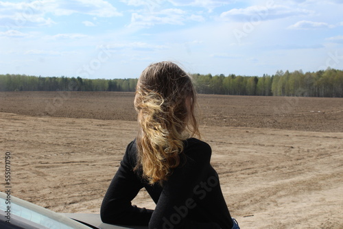 a girl with long blonde hair that flutters in the wind is leaning on a car and looking out into the field