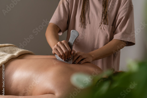 Hands of the masseur are close-up using a metal massage tool to massage the back of a woman lying on a couch in a cozy office. Health concept  body care  skin care  wellness 