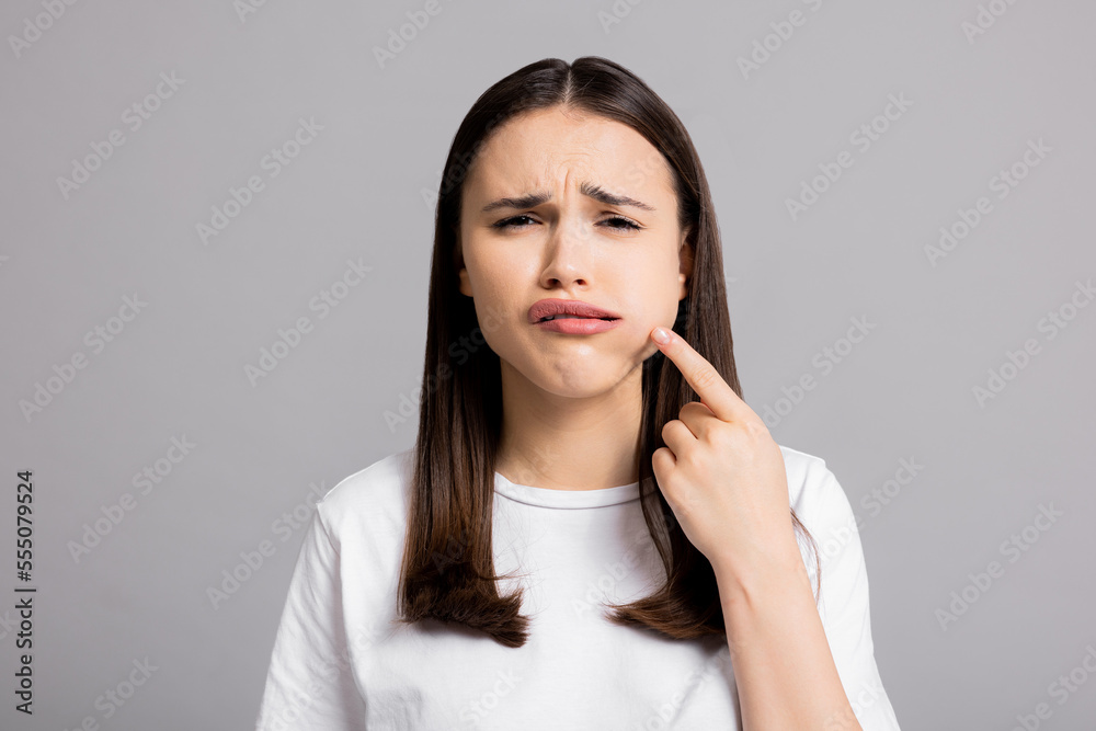 Crying tired sad woman suffers from painful toothache dental illness pointing on teeth standing on grey background in studio isolated wearing basic white t-shirt. Crying tired sad woman suffers from painful toothache dental illness pointing on teeth standing on grey background in studio isolated wearing basic white t-shirt.