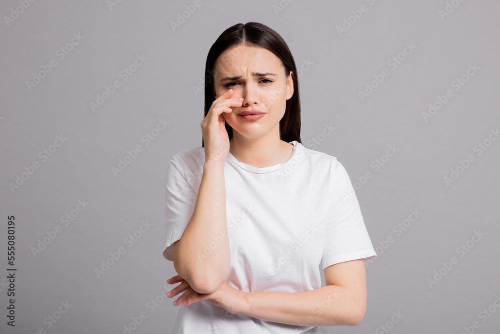 Cried sad young woman wearing white casual t-shirt standing on grey background in studio isolated. Girl suffering from abuse domestic volatile desperate woman. Cried sad young woman wearing white casual t-shirt standing on grey background in studio isolated. Girl suffering from abuse domestic volatile desperate woman.