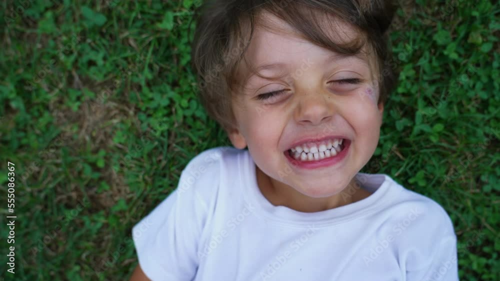 Happy child laughing close up face. Male kid laid on grass smiling. Real life laugh and smile. Authentic joy expression Happy child laughing close up face. Male kid laid on grass smiling. Real life laugh and smile. Authentic joy expression