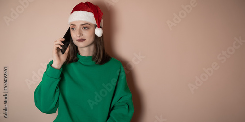 a girl in a New Year's hat is talking on the phone on a plain background, holding a phone in her hand, ordering dinner, the concept of New Year's weekend 2023, happy christmas, 2023, holidays