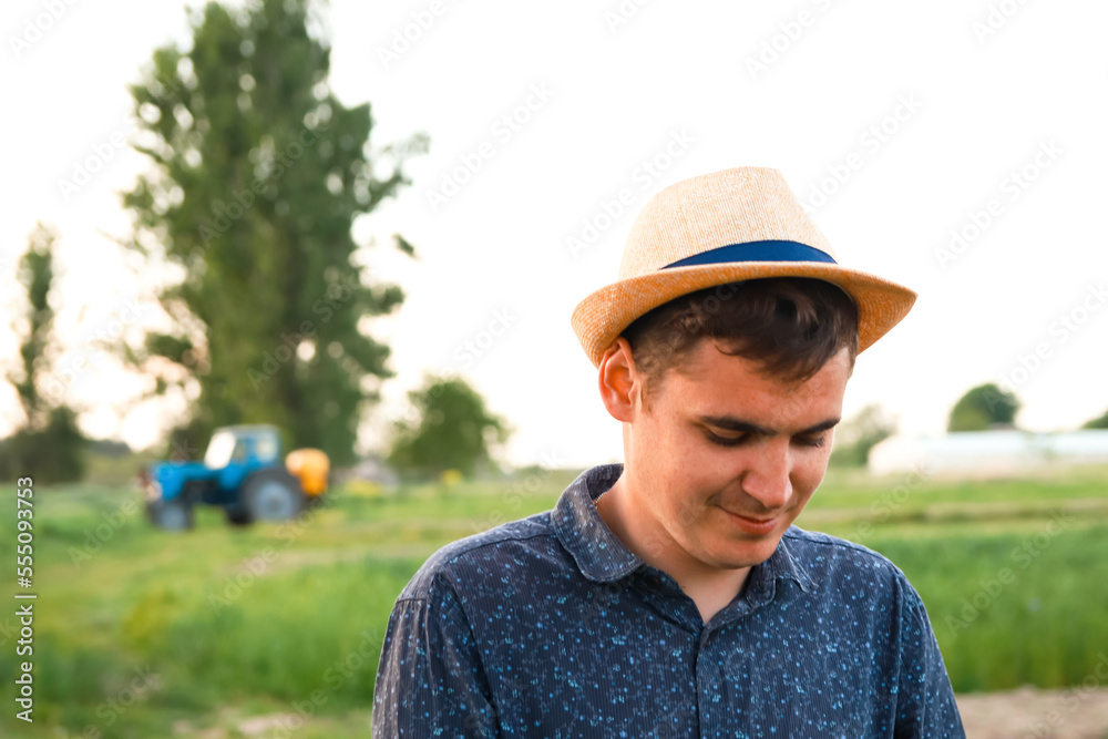 Defocus farmer in hat working on field with greenhouse outdoor. Portrait of young Caucasian handsome happy man farmer standing in field and smiling. Tractor on background. Out of focus