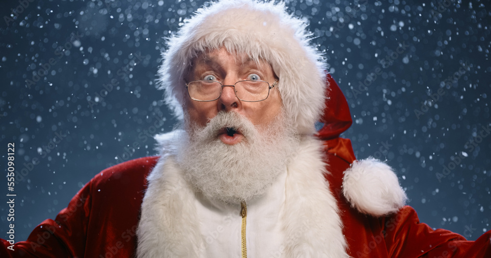 Santa Clause looking at camera when suddenly getting surprised and throwing his arms wide, isolated on snowy blue background - christmas spirit concept close up Santa Clause looking at camera when suddenly getting surprised and throwing his arms wide, isolated on snowy blue background - christmas spirit concept close up