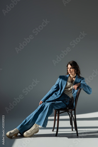 full length of stylish woman in blue suit and boots looking away on chair on grey background with lighting and shadows