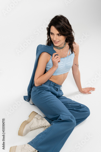 happy brunette woman in pants and boots sitting with blue jacket and looking away on grey