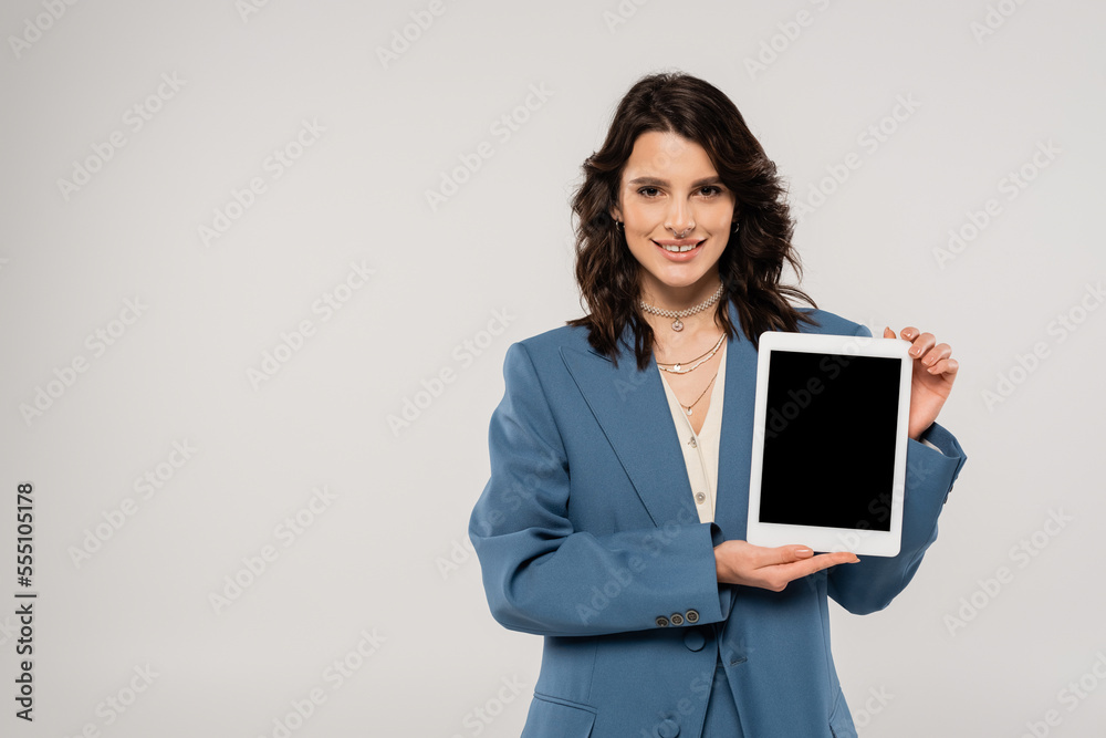 happy brunette woman in blue jacket smiling at camera and showing digital tablet with blank screen isolated on grey happy brunette woman in blue jacket smiling at camera and showing digital tablet with blank screen isolated on grey