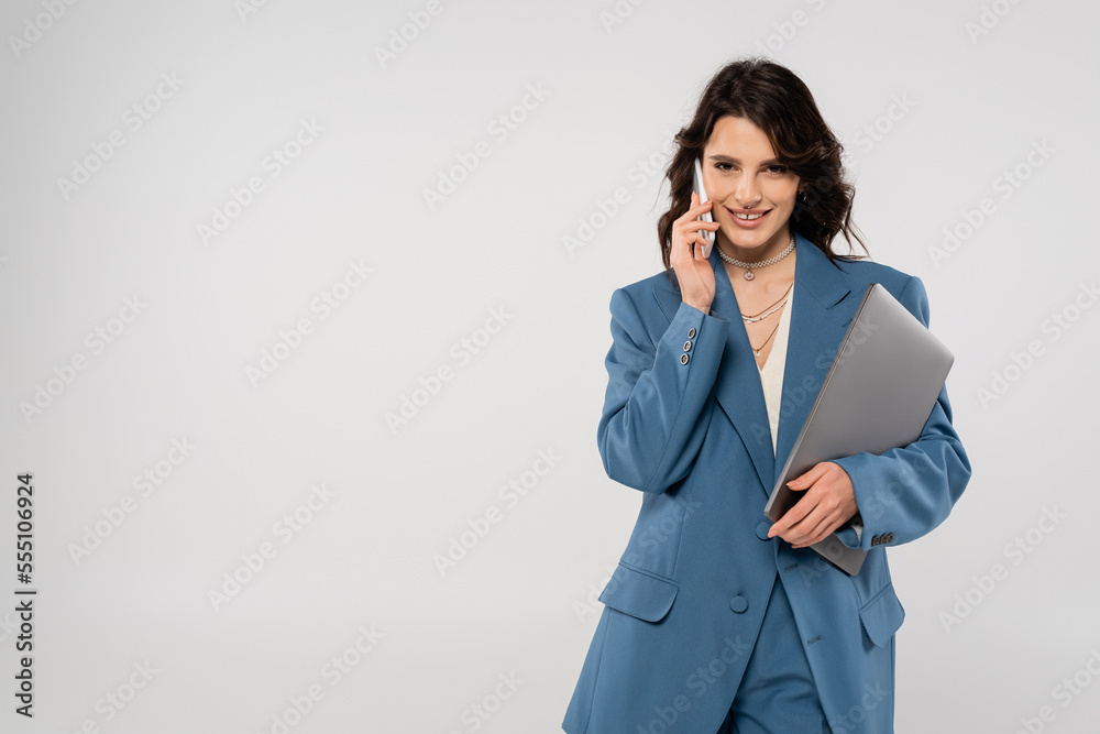 smiling brunette woman in blue blazer talking on cellphone and holding laptop isolated on grey smiling brunette woman in blue blazer talking on cellphone and holding laptop isolated on grey