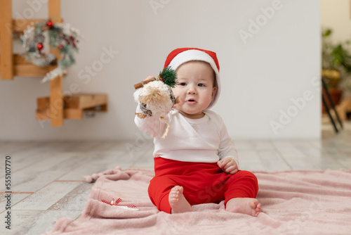 A 9-month-old Asian baby in a Christmas costume plays with a New year toy on a blanket laid out on the floor and looks into the camera with her eyes wide open and laughs
