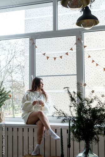 Woman in warm white winter sweater sitting at windowsill at home at christmas eve holding cup with marshmallows