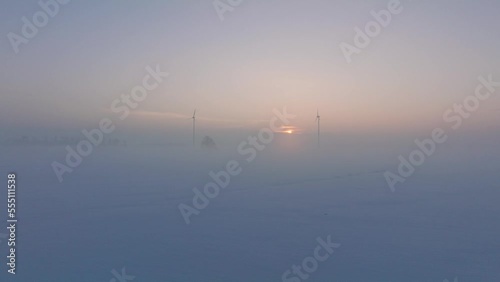 Aerial view of wind turbines generating renewable energy in the wind farm, snow filled countryside landscape with fog, sunny winter evening with golden hour light, wide drone shot moving forward low photo