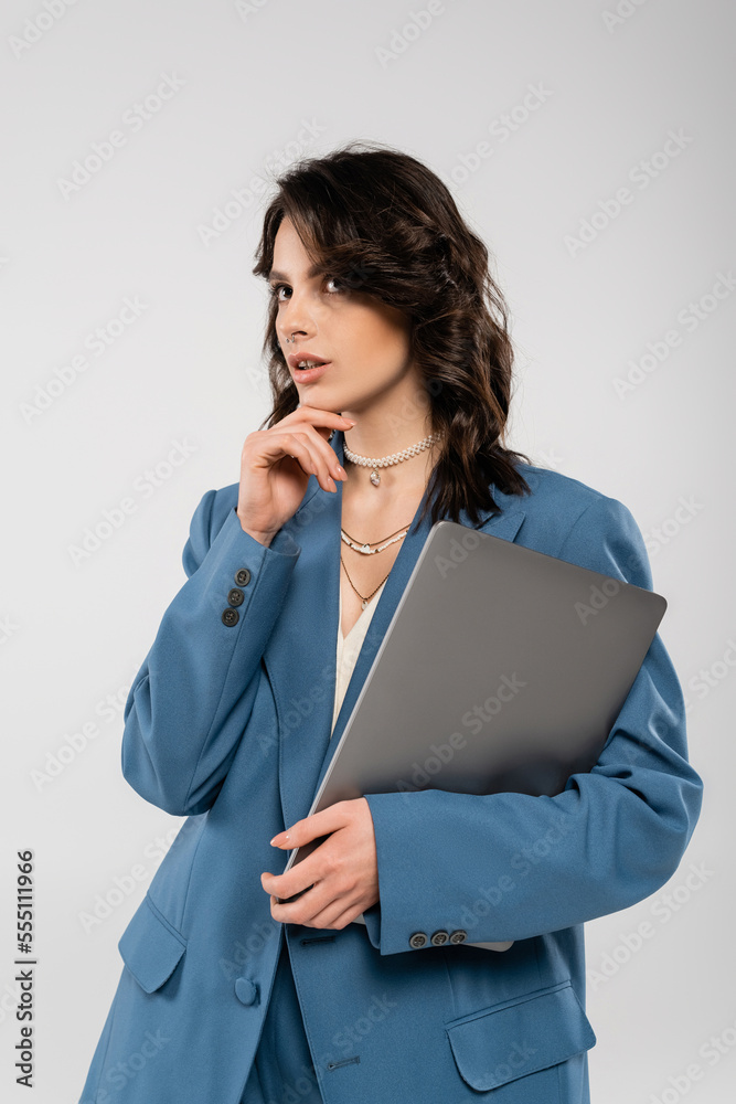 thoughtful brunette woman in blue jacket holding laptop and looking away isolated on grey thoughtful brunette woman in blue jacket holding laptop and looking away isolated on grey