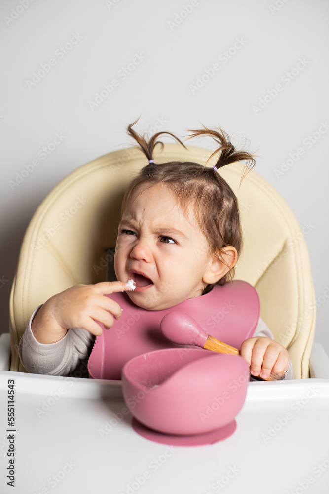 Small cute little toddler brunette caucasian girl with two tails eating by herself with a spoon and finger sitting in baby chair, dissatisfied with the food; self-feeding concept Small cute little toddler brunette caucasian girl with two tails eating by herself with a spoon and finger sitting in baby chair, dissatisfied with the food; self-feeding concept