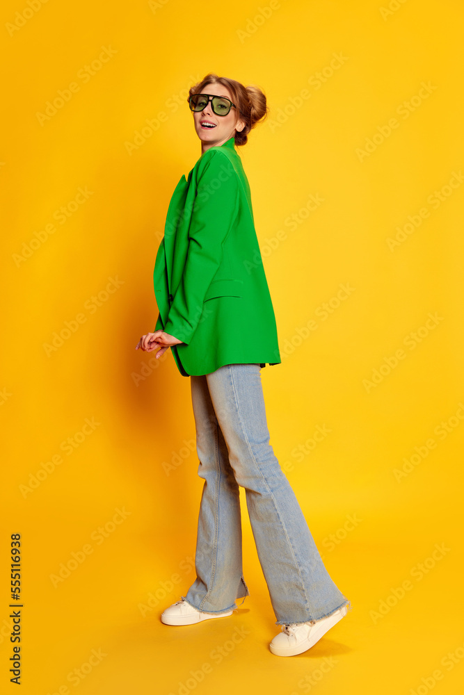 Fototapeta premium Portrait of young girl in bright green jacket posing over yellow studio background. Stylish, fashionable youth