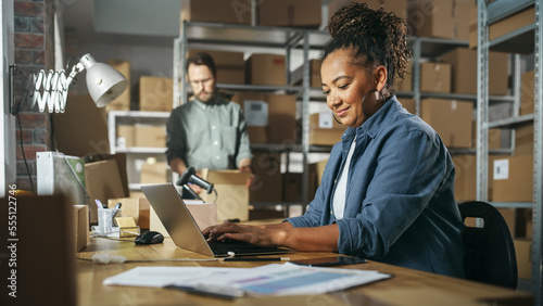 Diverse Male and Female Warehouse Inventory Managers Talking, Using Laptop Computer and Checking Retail Stock. Rows of Shelves Full of Cardboard Box Packages in the Background.