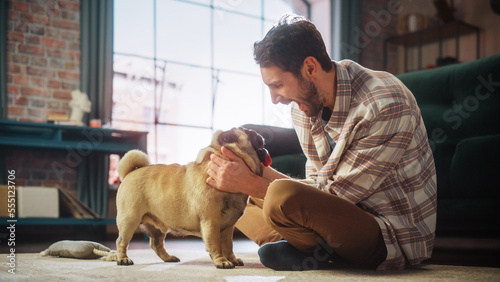 Happy Man Cuddles His Adorable Little Pug, Gives it Snacks at Home. Handsome Man Plays with His Dog, Best Friend, Scratches Super Happy Doggy, Have Fun in the Living Room