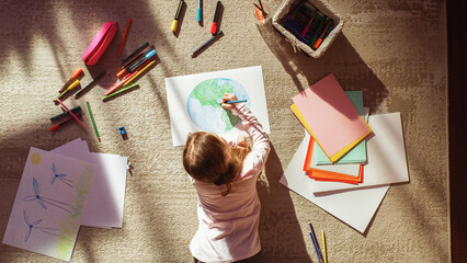 Top View: Little Girl Drawing Our Beautiful Green Planet Earth. Child Having Fun at Home on the Floor, Imagining Our Planet as a Happy Place with Clean, Sustainable Living. Cozy Sunny Day.