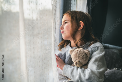 Fotografia a pensive cute girl sits and looks out the window, hugging a teddy bear