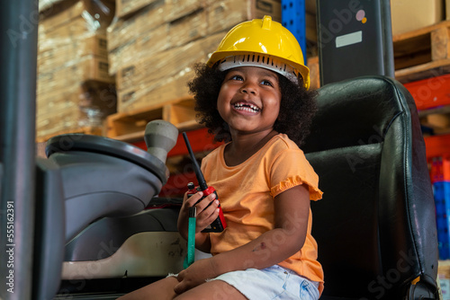 three-year-old African-American girl in an engineer's helmet smiling happily drives a forklift as an engineer in a factory.