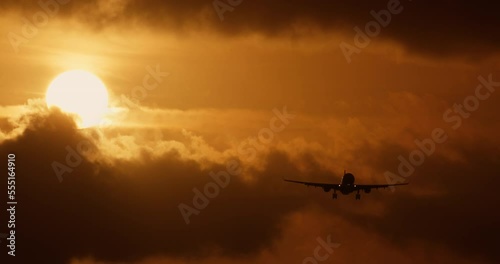 Magical and magnificent view of flying plane, behind which orange disk of warm summer sun peeks out from behind clouds. Incredible and epic sunset, cloudy orange sky with flying silhouette of airplane