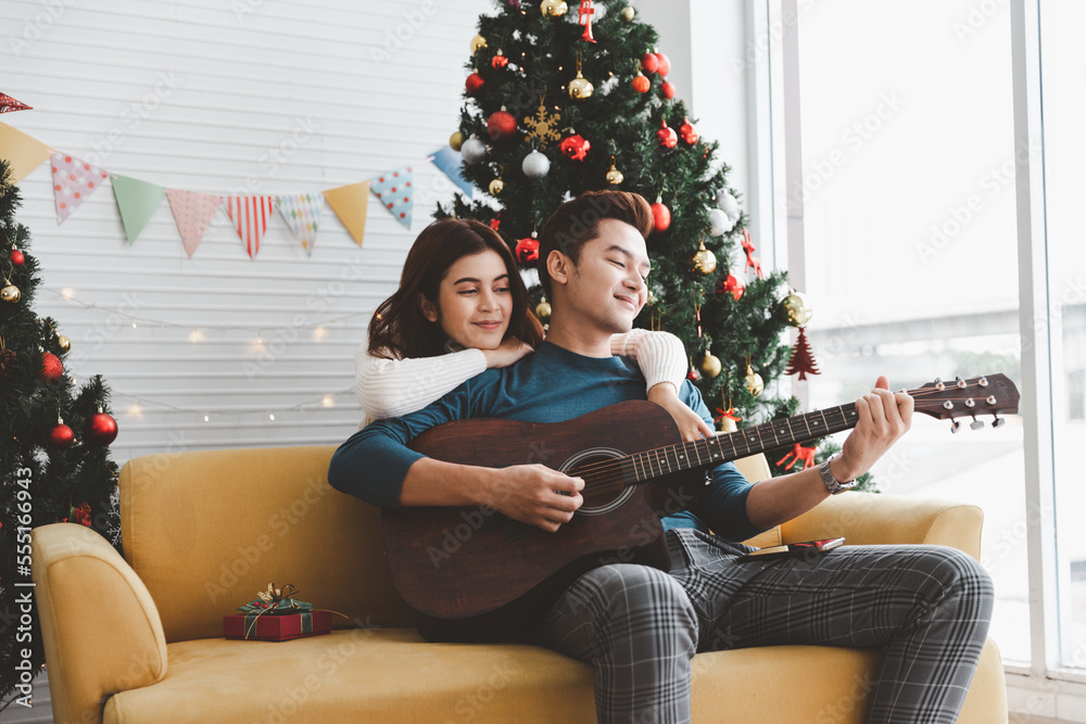 A man playing guitar to woman lover during celebrateing christmas holiday together in living room with christmas tree ornament decoration A man playing guitar to woman lover during celebrateing christmas holiday together in living room with christmas tree ornament decoration