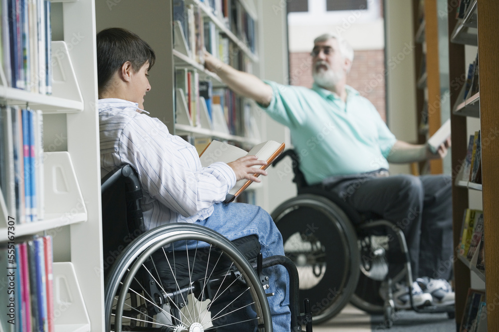 Middle-aged man with Muscular Dystrophy picking a book from a bookshelf with a young woman reading a book in a library Middle-aged man with Muscular Dystrophy picking a book from a bookshelf with a young woman reading a book in a library