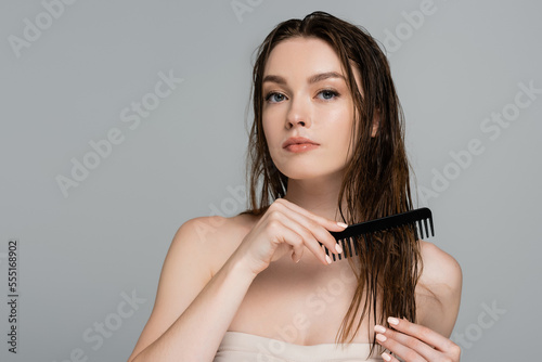pretty young woman with bare shoulders brushing wet hair with comb isolated on grey