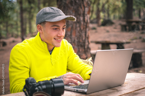 Business on fresh air for a beautiful young freelancer working on laptop sitting outdoors in the forest. Hipster boy traveler working distantly while enjoying nature mountain during vacations © luciano