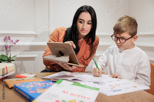 Una donna mora aiuta un bambino biondo a fare i compiti  seduti in una sala elegante con l'aiuto di un tablet