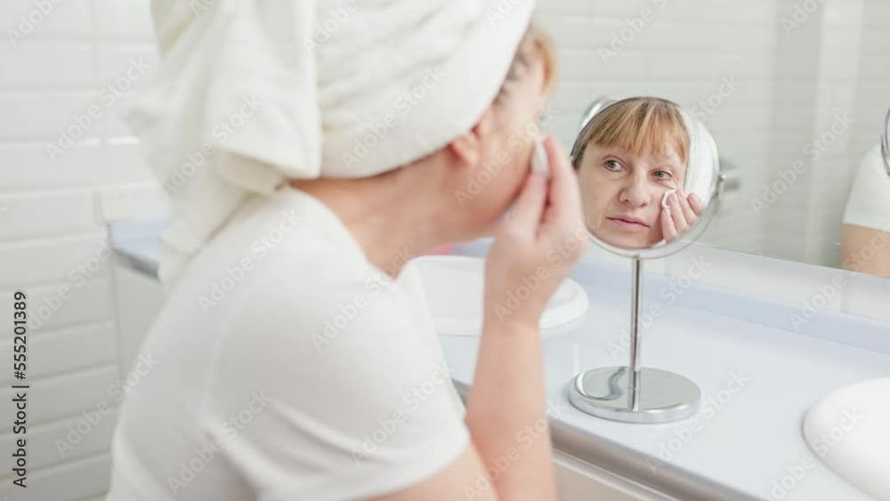 Gorgeous mid age older adult 50 years old blonde woman wears bathrobe in bathroom applying nourishing antiage face skin care cream treatment, looking at mirror doing daily morning beauty routine. Gorgeous mid age older adult 50 years old blonde woman wears bathrobe in bathroom applying nourishing antiage face skin care cream treatment, looking at mirror doing daily morning beauty routine.