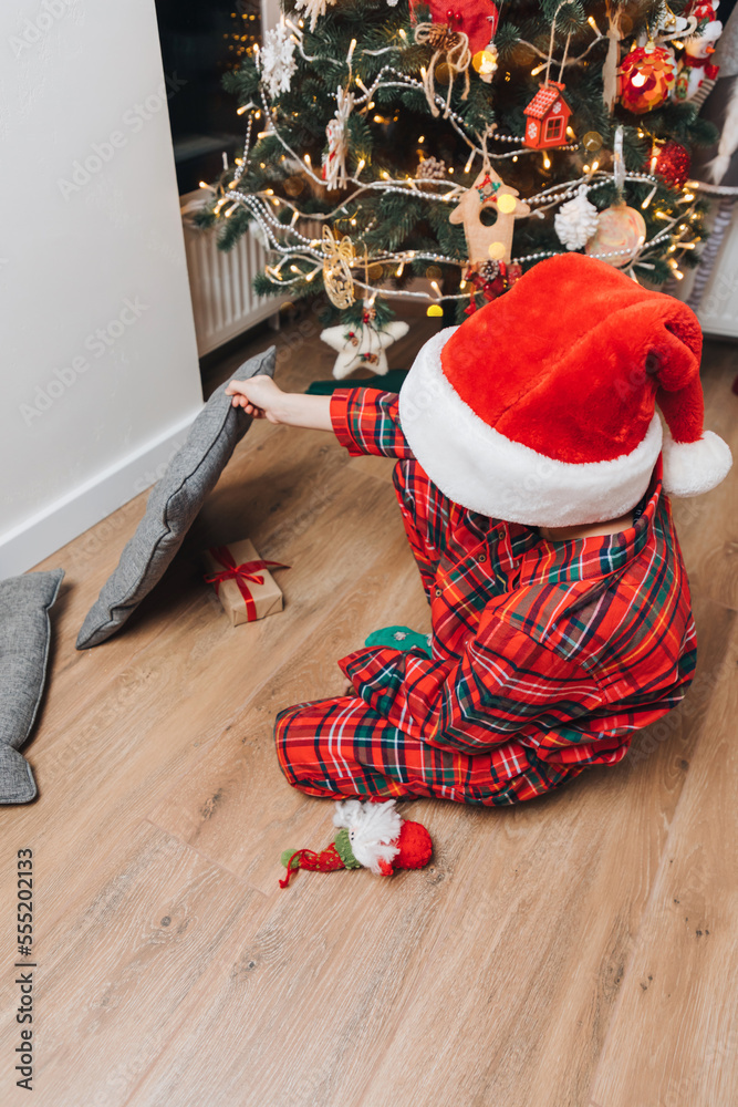 Obraz premium A boy in red pajamas near the Christmas tree is looking for a gift. Christmas at home. Family comfort. Front view