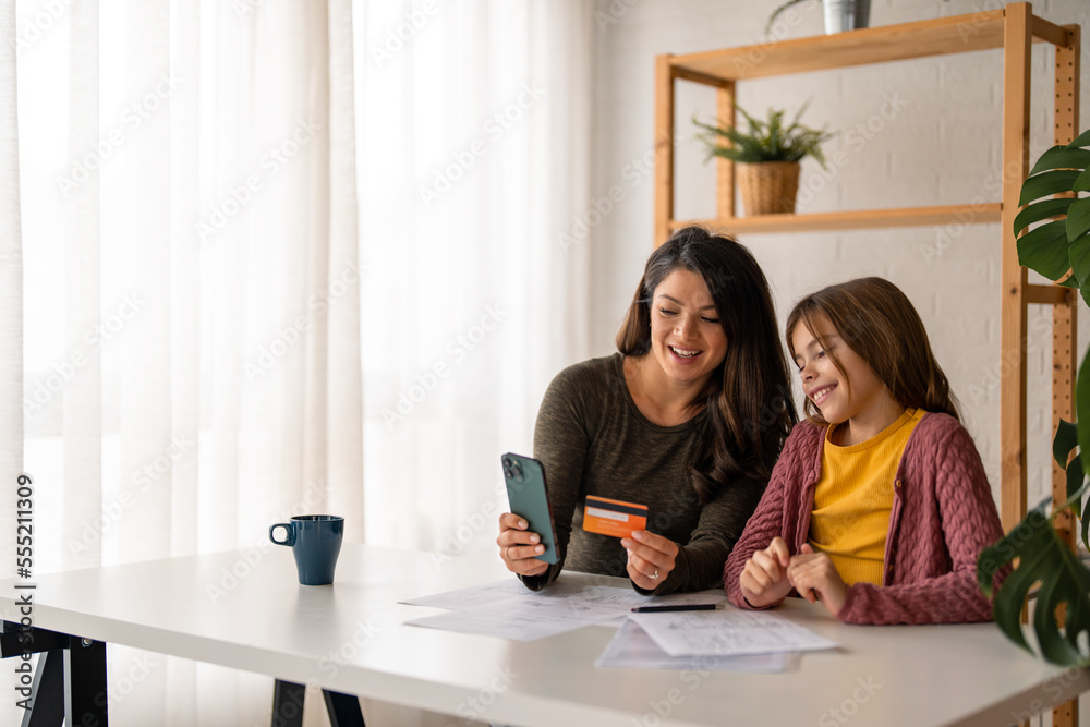 Young mom with school age daughter making purchases in popular internet store from home. Smiling woman holding credit card for online payments. Young mom with school age daughter making purchases in popular internet store from home. Smiling woman holding credit card for online payments.