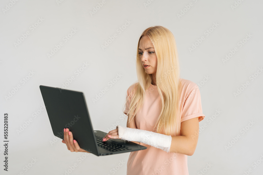 Portrait of scared blonde young woman with broken arm wrapped in plaster bandage holding laptop and working typing, standing on white isolated background. Concept of insurance and healthcare. Portrait of scared blonde young woman with broken arm wrapped in plaster bandage holding laptop and working typing, standing on white isolated background. Concept of insurance and healthcare.