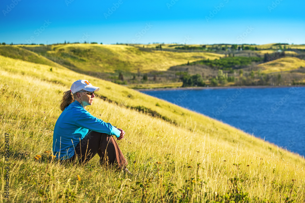 Female sitting on grassy hillside overlooking lake and rolling hills in the background with blue sky, Waterton Lakes National Park; Waterton, Alberta, Canada Female sitting on grassy hillside overlooking lake and rolling hills in the background with blue sky, Waterton Lakes National Park; Waterton, Alberta, Canada