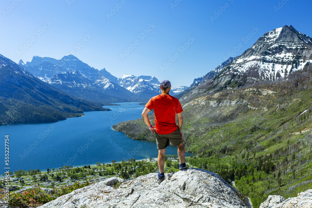 A male hiker standing on top of a rock cliff overlooking the mountains and a lake in the distance with blue sky, Waterton Lakes National Park; Waterton, Alberta, Canada