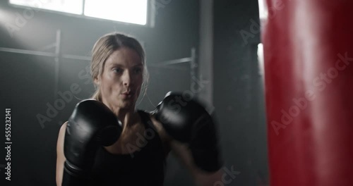 Beautiful energetic thin blonde young female box fighter beating punching bag training working out in gym. Focused confident serious professional woman boxer punching in gloves and top. photo