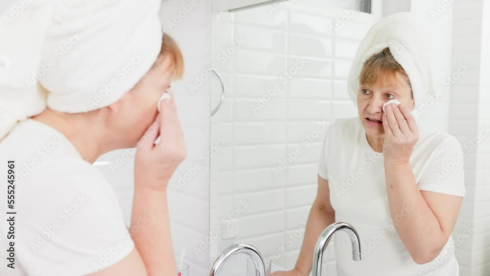 Gorgeous mid age older adult 50 years old blonde woman wears bathrobe in bathroom applying nourishing antiage face skin care cream treatment, looking at mirror doing daily morning beauty routine. Gorgeous mid age older adult 50 years old blonde woman wears bathrobe in bathroom applying nourishing antiage face skin care cream treatment, looking at mirror doing daily morning beauty routine.