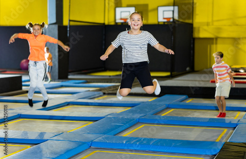 Joyful school-age girl in casual clothes bouncing on trampolines indoors in leisure sport center for kids