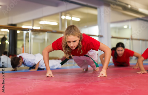 Sporty preteen girl doing push-ups on gym floor during mugendo group training