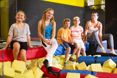 Cheerful friends, preteen girls and boys, enjoying spending time together in indoor inflatable leisure park, resting on padded log in pool with soft yellow foam cubes