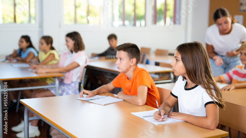 Interested intelligent preteen girl studying in classroom with classmates, listening attentively to teacher and writing in notebook photo