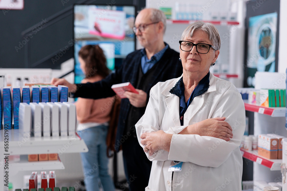 Portrait of senior pharmacist looking at camera while standing with arm crossed in pharmacy, offering support to clients. Customers looking at drugstore shelves buying pills and vitamins Portrait of senior pharmacist looking at camera while standing with arm crossed in pharmacy, offering support to clients. Customers looking at drugstore shelves buying pills and vitamins
