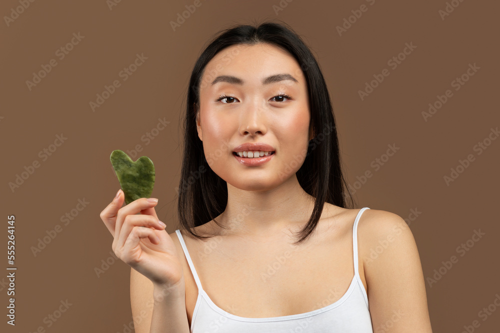 Women beauty trends. Young asian lady holding gua sha lymphatic drainage facial self massager, brown studio background Women beauty trends. Young asian lady holding gua sha lymphatic drainage facial self massager, brown studio background