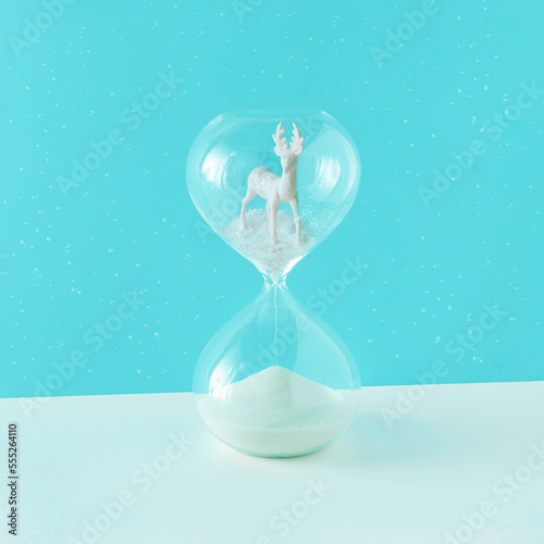 2023. White reindeer standing in the snow in the hourglass on isolated snowy pastel blue background. Minimal abstract aesthetic Xmas greeting card. New Year holiday decoration concept.